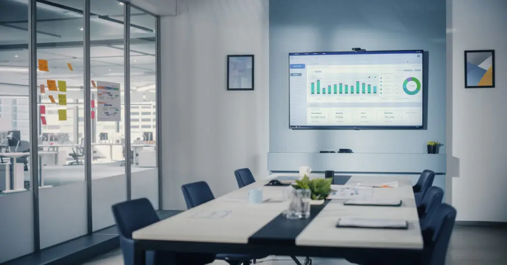 Modern Empty Meeting Room with Big Conference Table with Various Documents and Laptops on it, on the Wall Big TV with Big Data Analysis